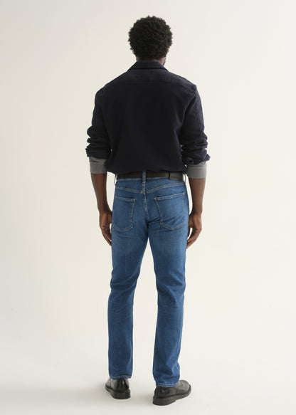 Person wearing medium blue straight leg jeans with a navy long sleeve collared pullover and black shoes, back full body view on a light grey background.