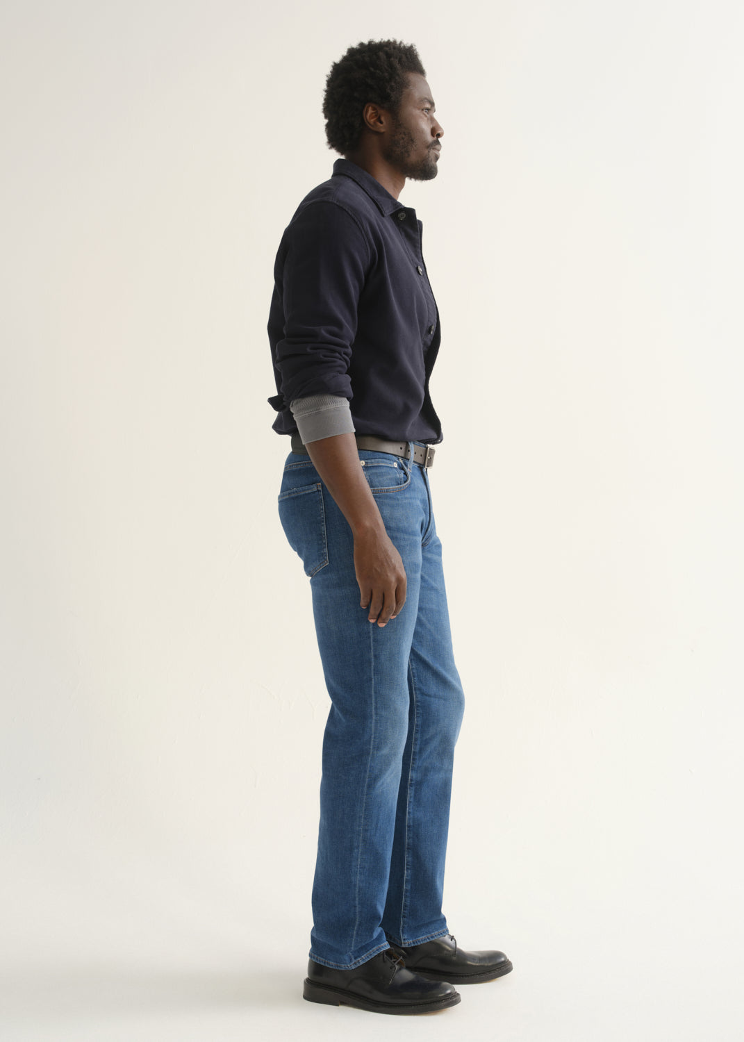 Person wearing medium blue straight leg jeans with a navy long sleeve collared pullover and black shoes, side full body view on a light grey background.