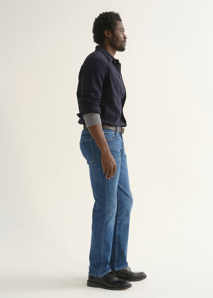 Person wearing medium blue straight leg jeans with a navy long sleeve collared pullover and black shoes, side full body view on a light grey background.