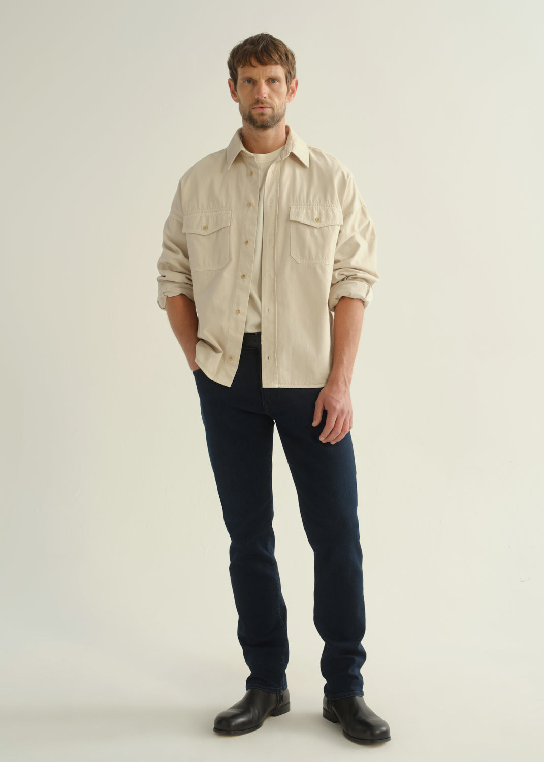 Person wearing a cream button-front shirt, dark indigo jeans, and black boots, front full body view on a light grey background.