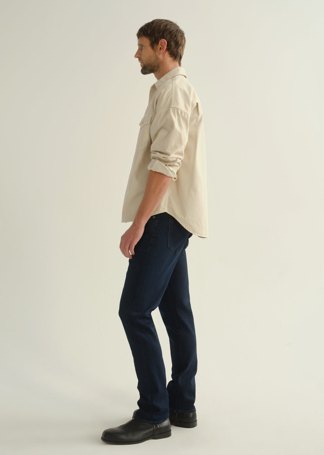 Person wearing a cream button-front shirt, dark indigo jeans, and black boots, side full body view on a light grey background.