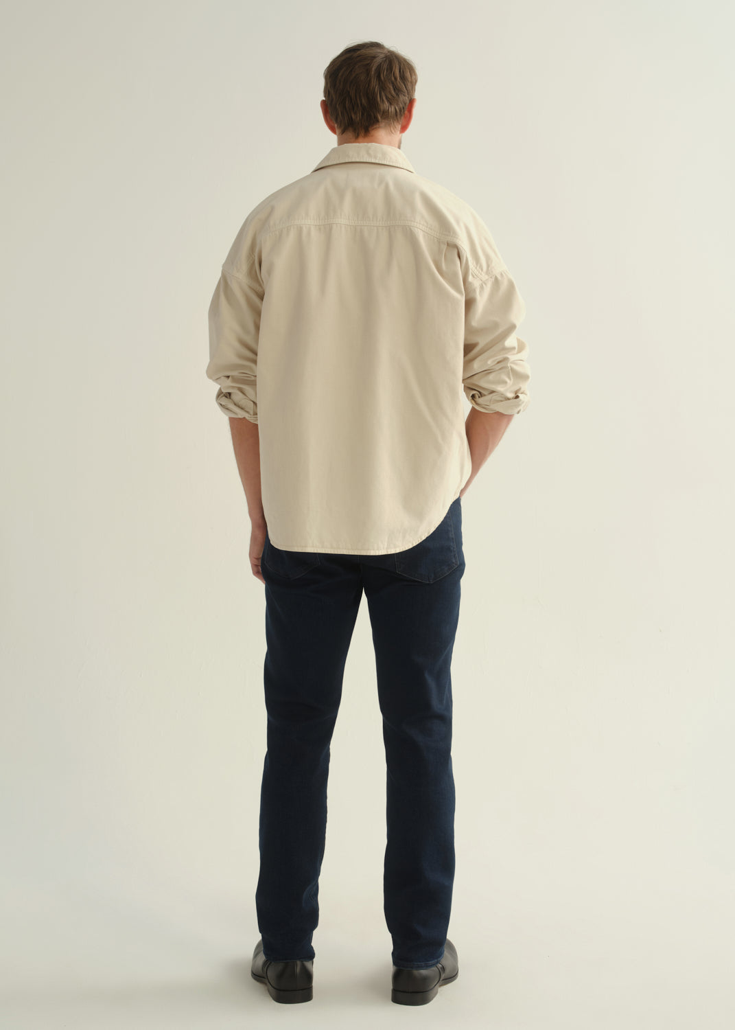Person wearing a cream button-front shirt, dark indigo jeans, and black boots, back full body view on a light grey background.
