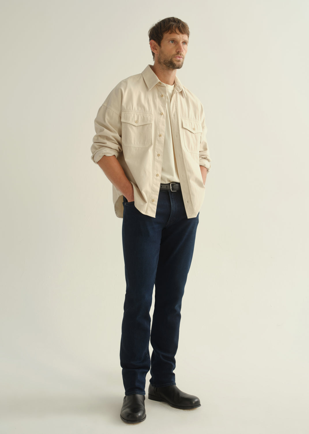 Person wearing a cream button-front shirt, dark indigo jeans, and black boots, angled front full body view on a light grey background.