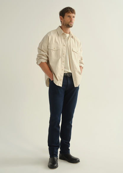 Person wearing a cream button-front shirt, dark indigo jeans, and black boots, angled front full body view on a light grey background.