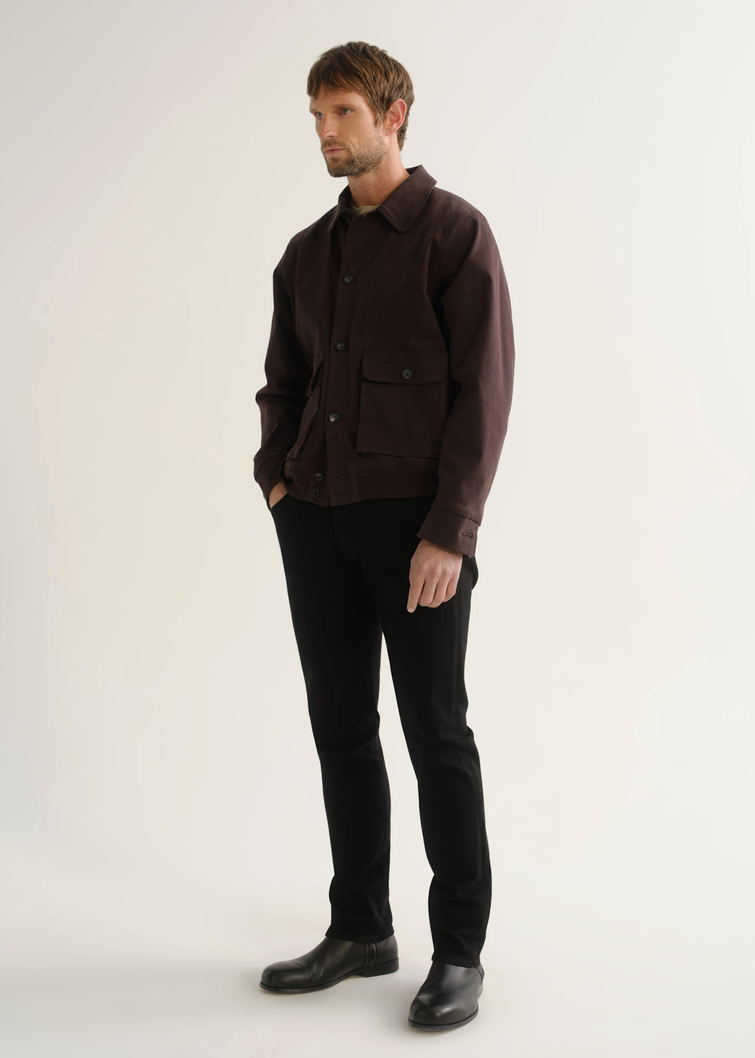 Person wearing a dark brown jacket, black pants, and black boots, angled front full body view on a light grey background.
