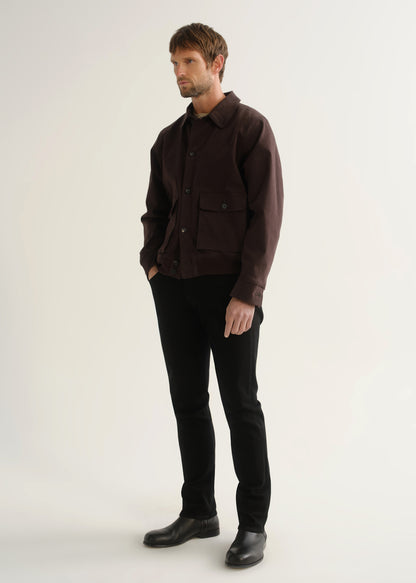 Person wearing a dark brown jacket, black pants, and black boots, angled front full body view on a light grey background.