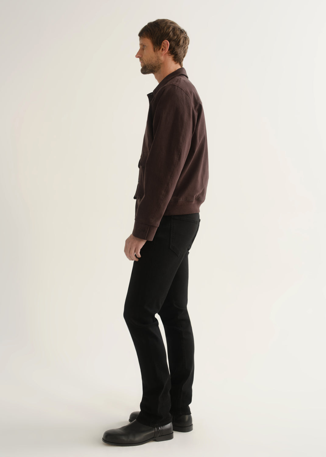 Person wearing a dark brown jacket, black pants, and black boots, side full body view on a light grey background.