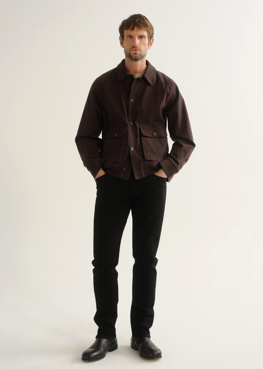 Person wearing a dark brown jacket, black pants, and black boots, front full body view on a light grey background.