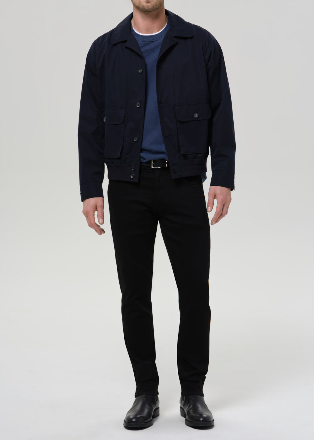 A man wearing a dark navy blue utility jacket over a blue t-shirt, black trousers, and black leather shoes, standing against a grey background.