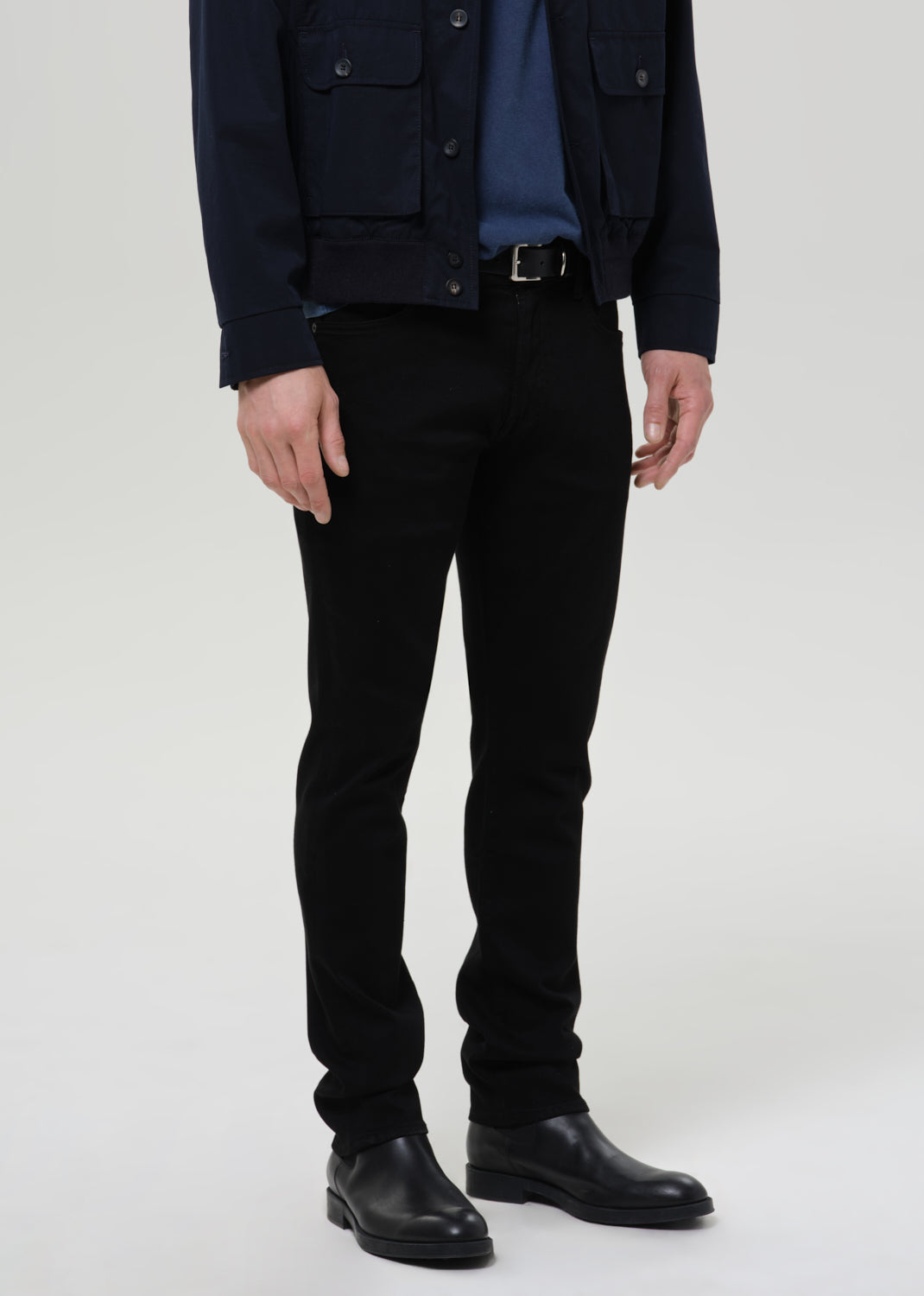 A model wearing slim-fit black trousers, styled with a navy blue jacket, a blue shirt, and black leather boots, photographed against a grey background.