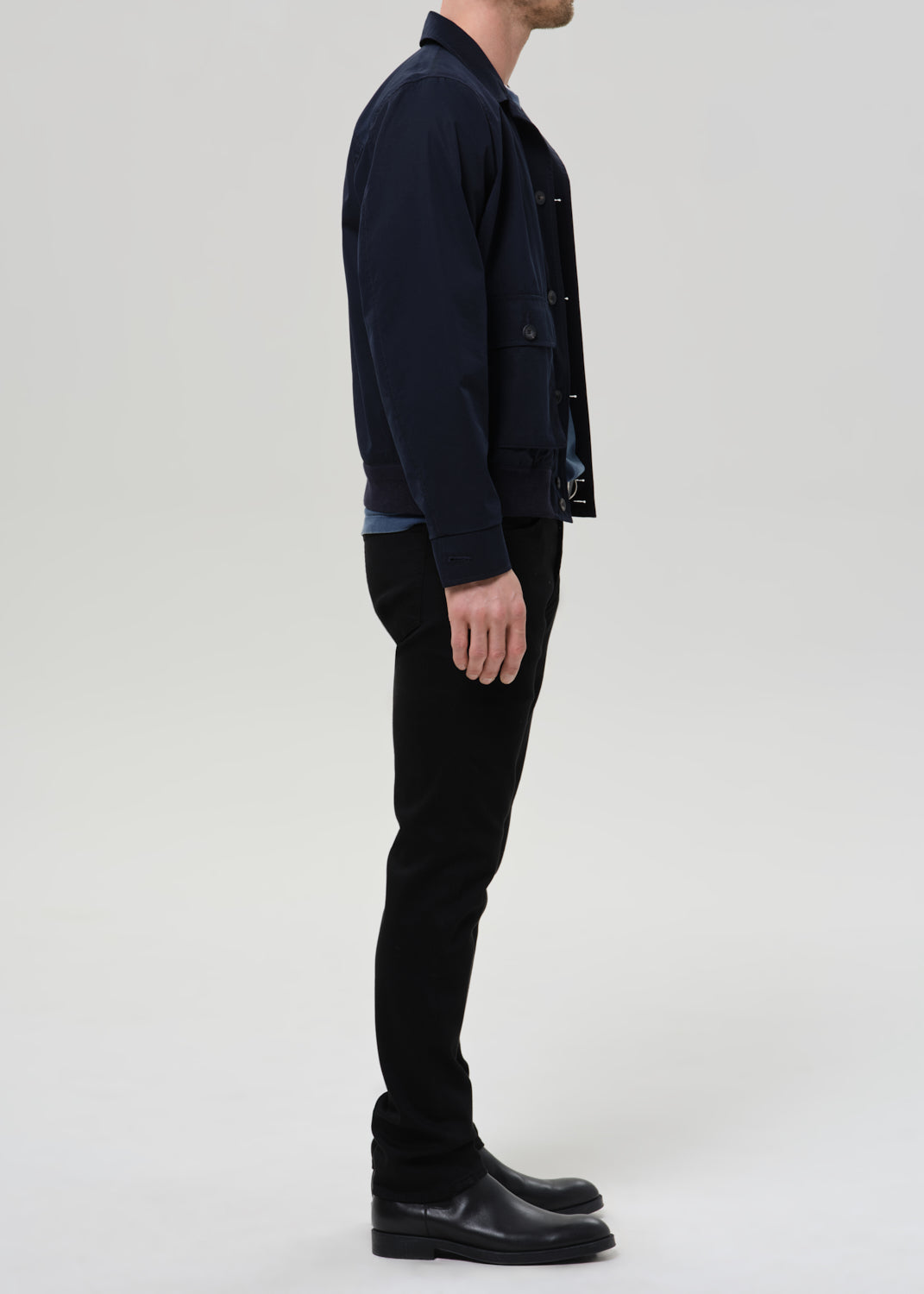 A man shown in profile wearing a dark navy jacket, black pants, and black leather boots against a grey background.