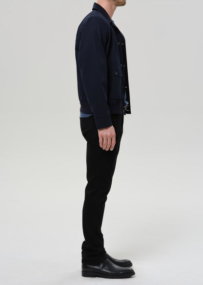 A man shown in profile wearing a dark navy jacket, black pants, and black leather boots against a grey background.