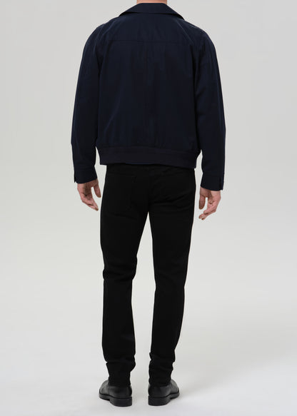 A man viewed from the back wearing a dark navy jacket, black pants, and black shoes, standing against a grey background.