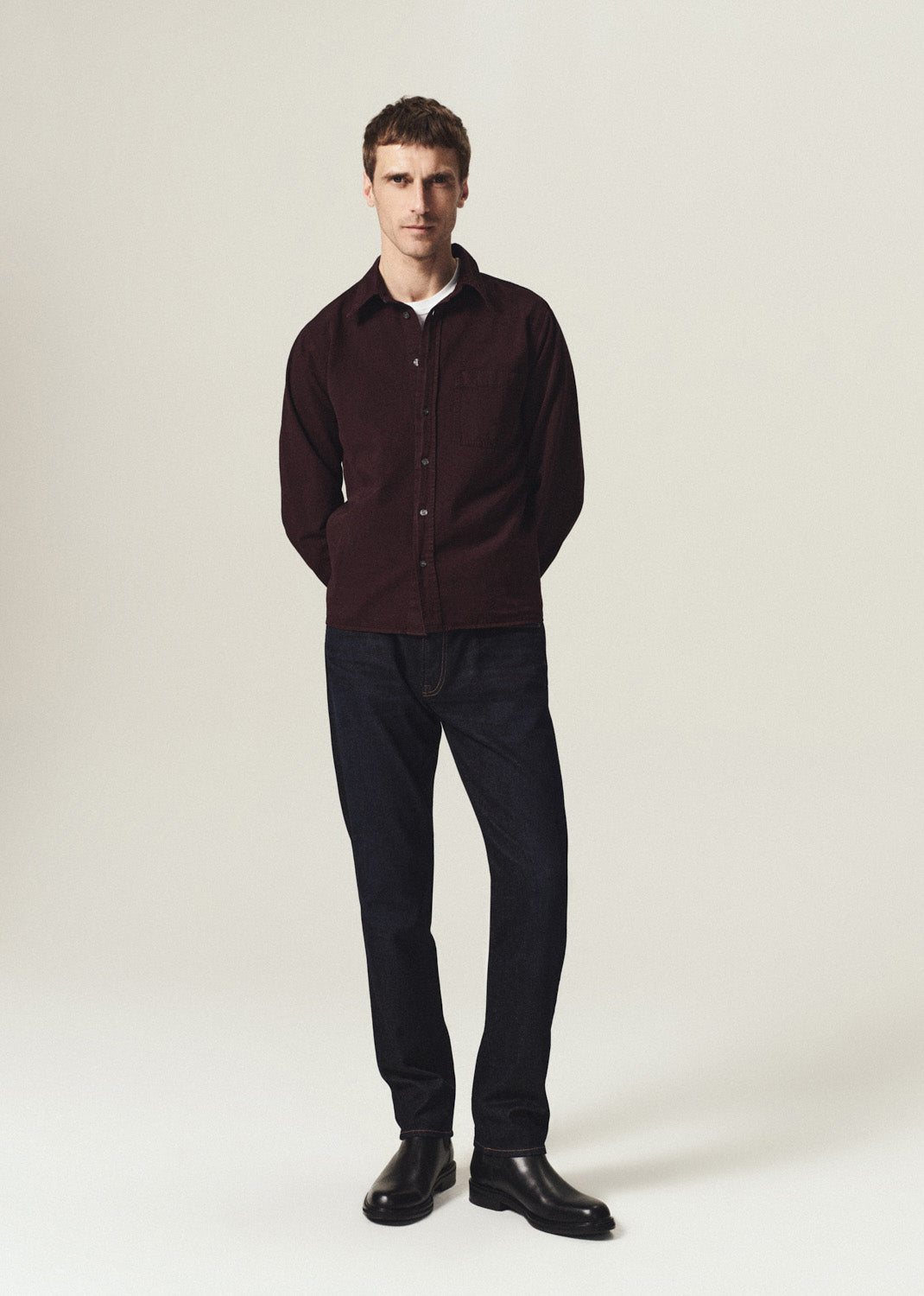 A man modeling a dark burgundy corduroy button-up shirt over a white undershirt, paired with dark trousers and black shoes, standing against a plain light beige studio background.
