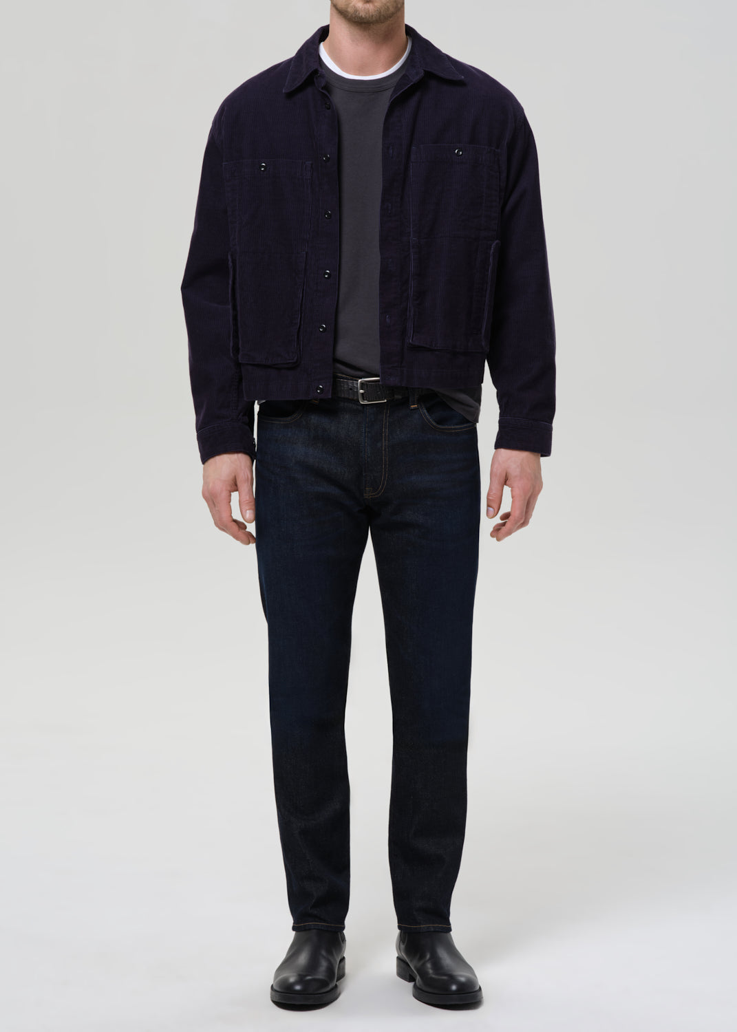 A man wearing a dark navy corduroy jacket over a dark gray t-shirt, dark blue jeans, and black leather boots, standing against a grey background.