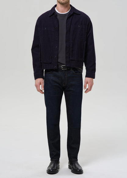 A man wearing a dark navy corduroy jacket over a dark gray t-shirt, dark blue jeans, and black leather boots, standing against a grey background.