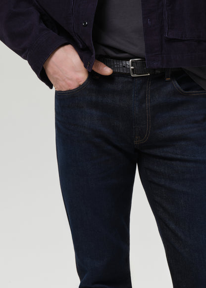 Close-up of a person wearing dark indigo denim jeans with a black belt, a dark gray t-shirt, and a navy overshirt, standing against a grey background.