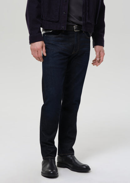 A person wearing slim-fit dark blue denim jeans, styled with a dark jacket, black shirt, belt, and black boots, photographed against a grey background.