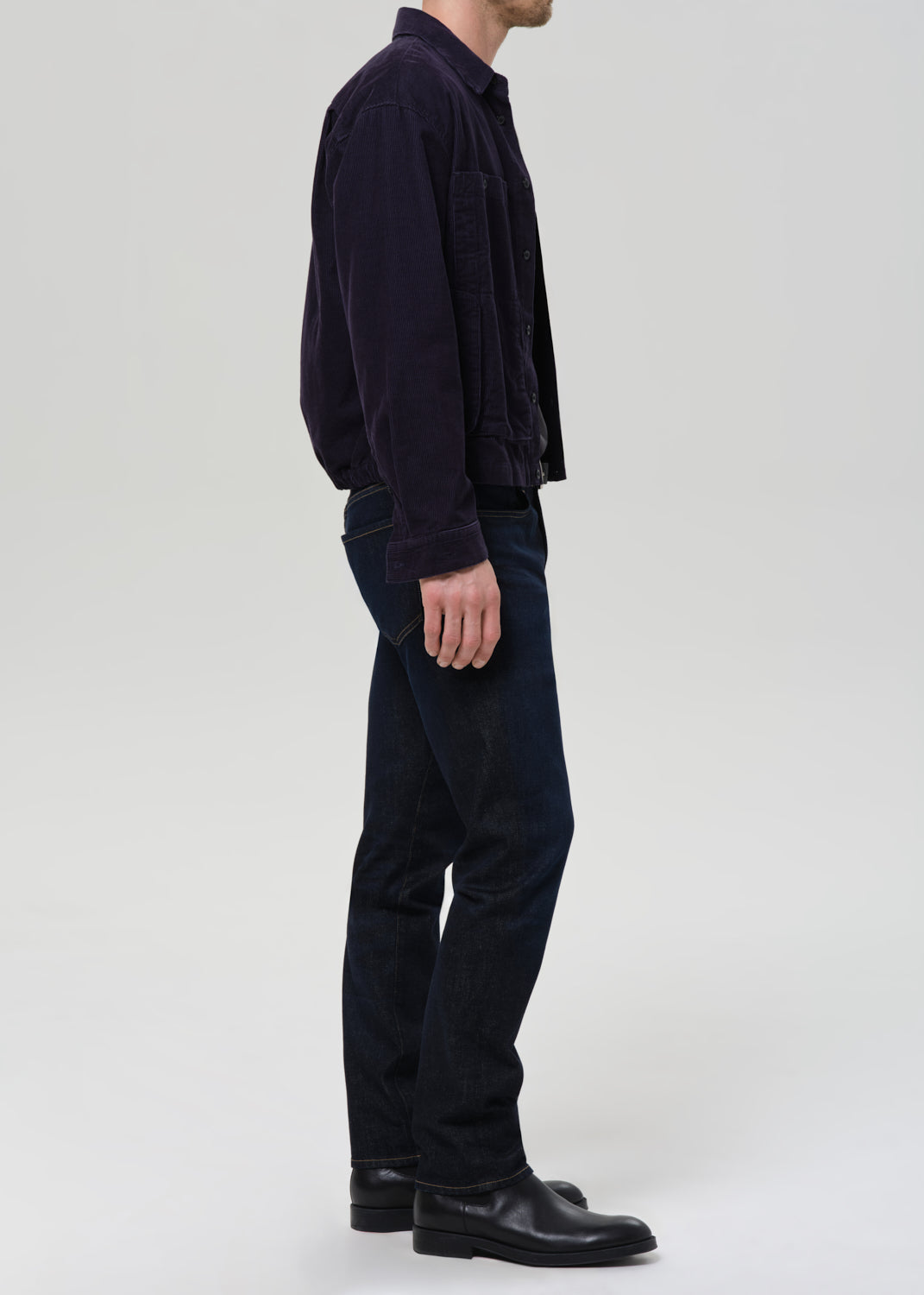 A man shown in profile wearing a dark navy corduroy jacket, dark blue jeans, and black leather shoes against a grey background.