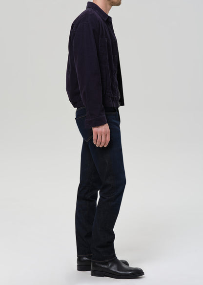 A man shown in profile wearing a dark navy corduroy jacket, dark blue jeans, and black leather shoes against a grey background.