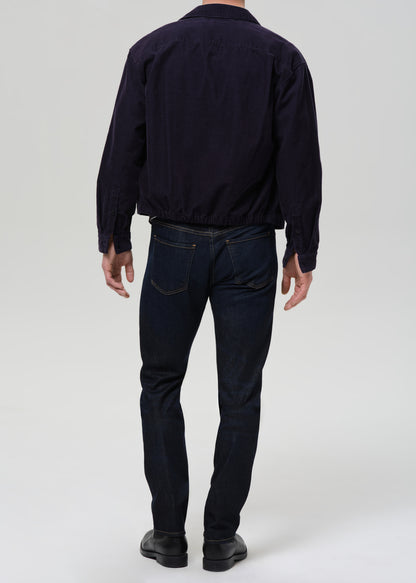 A man viewed from the back wearing a dark navy blue jacket and dark blue jeans, standing against a grey background.