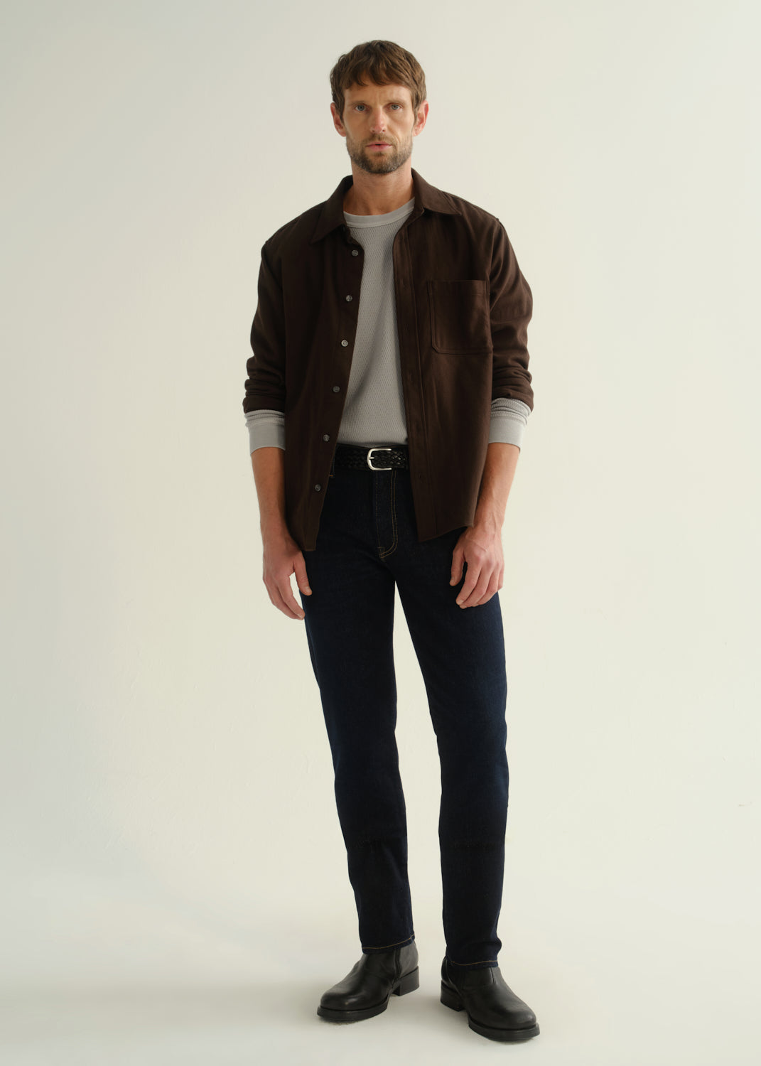 Person wearing a dark brown button-front shirt over a grey t-shirt, dark indigo jeans, and black boots, front full body view on a light grey background.