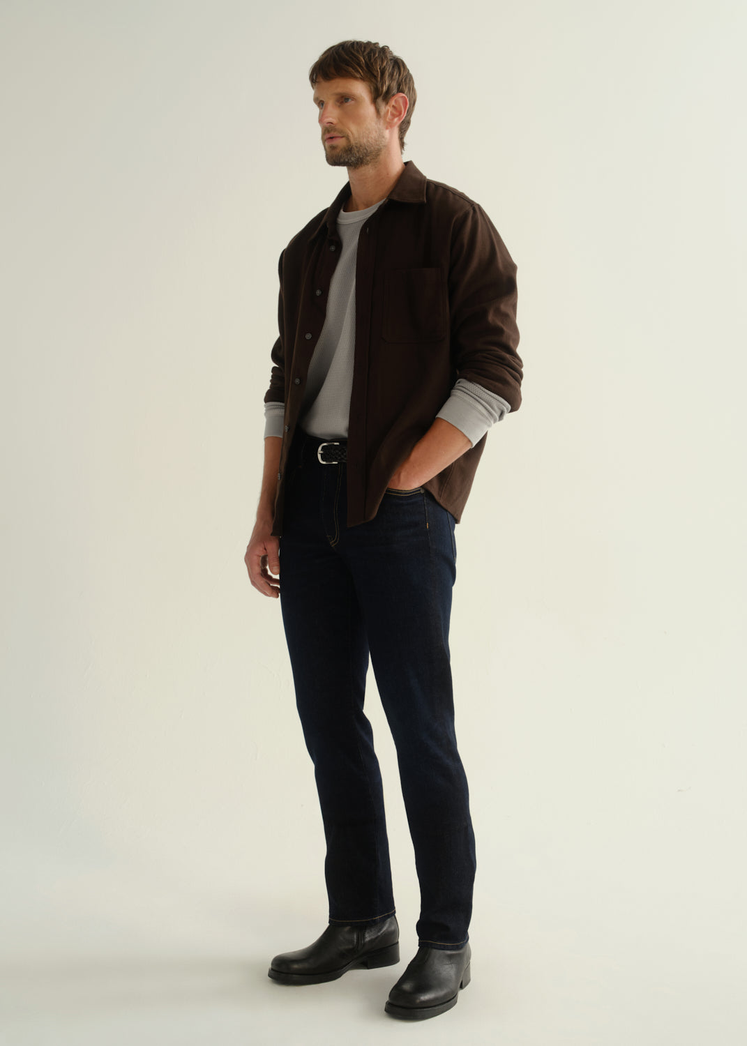 Person wearing a dark brown button-front shirt, dark indigo jeans, and black boots, side full body view on a light grey background.