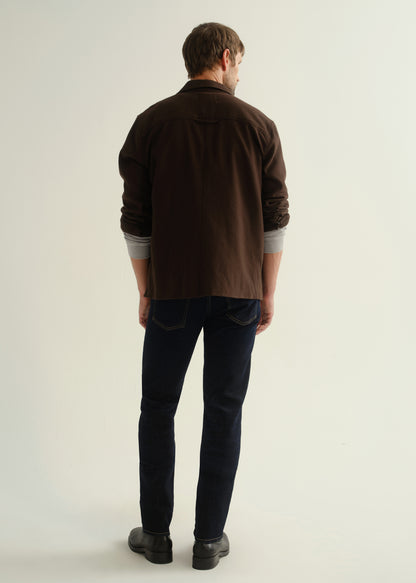 Person wearing a dark brown button-front shirt, dark indigo jeans, and black boots, back full body view on a light grey background.