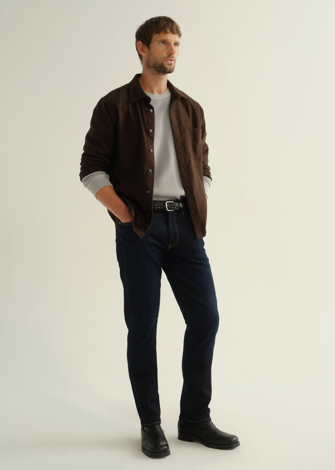Person wearing a dark brown button-front shirt over a grey t-shirt, dark indigo jeans, and black boots, angled front full body view on a light grey background.