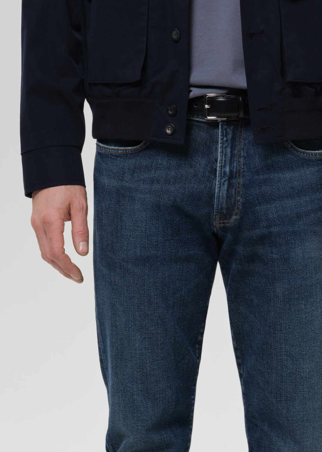 A close-up of a person wearing dark blue denim jeans, a black belt, a gray shirt, and a dark navy jacket, shown from the chest down against a grey background.