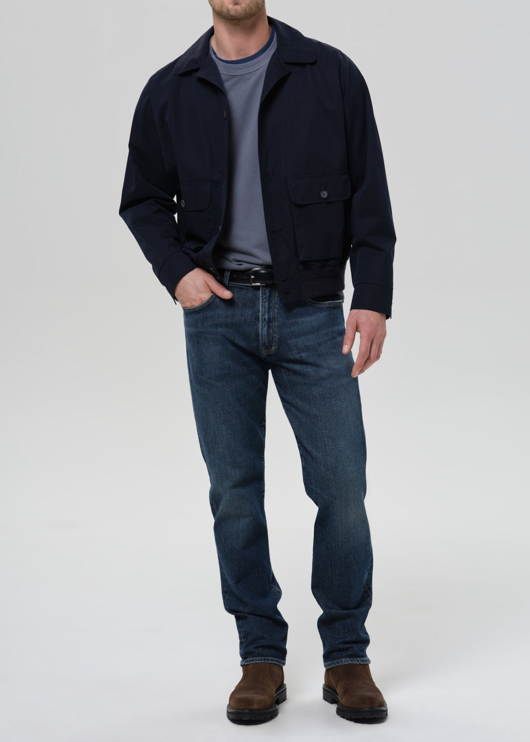 A man wearing a dark navy blue jacket over a gray t-shirt, blue jeans, and brown boots, standing against a grey background.