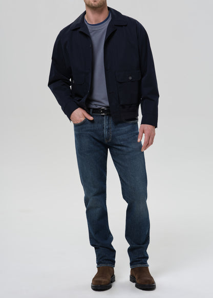A man wearing a dark navy blue jacket over a gray t-shirt, blue jeans, and brown boots, standing against a grey background.