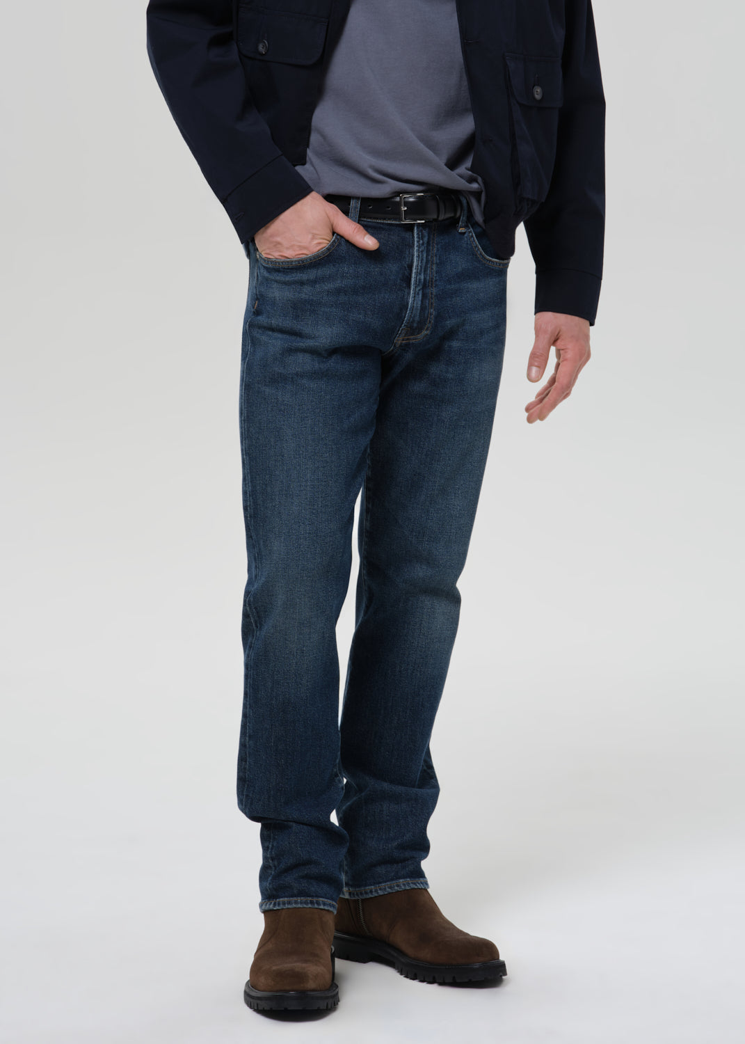 A person wearing dark blue slim-fit jeans, styled with a dark navy jacket, gray t-shirt, black belt, and brown suede boots, standing against a grey background.