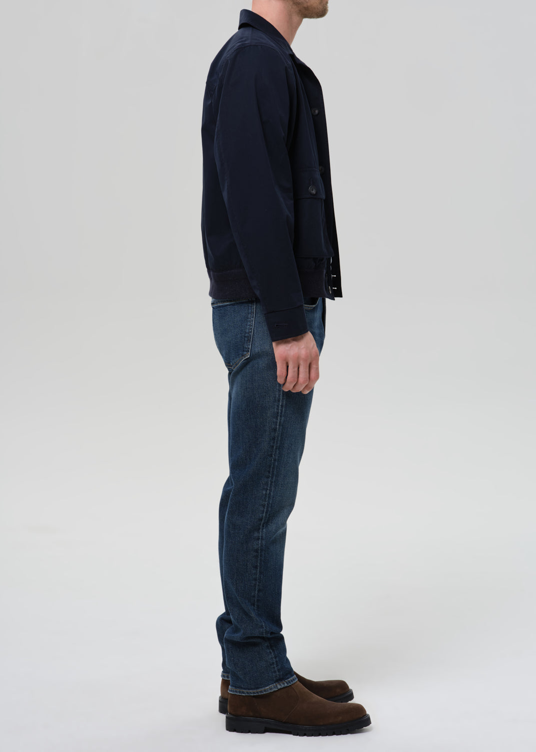 A man shown in profile wearing a dark navy jacket, blue jeans, and brown suede boots against a grey background.