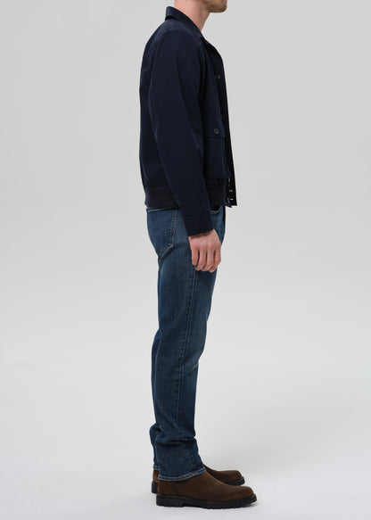 A man shown in profile wearing a dark navy jacket, blue jeans, and brown suede boots against a grey background.