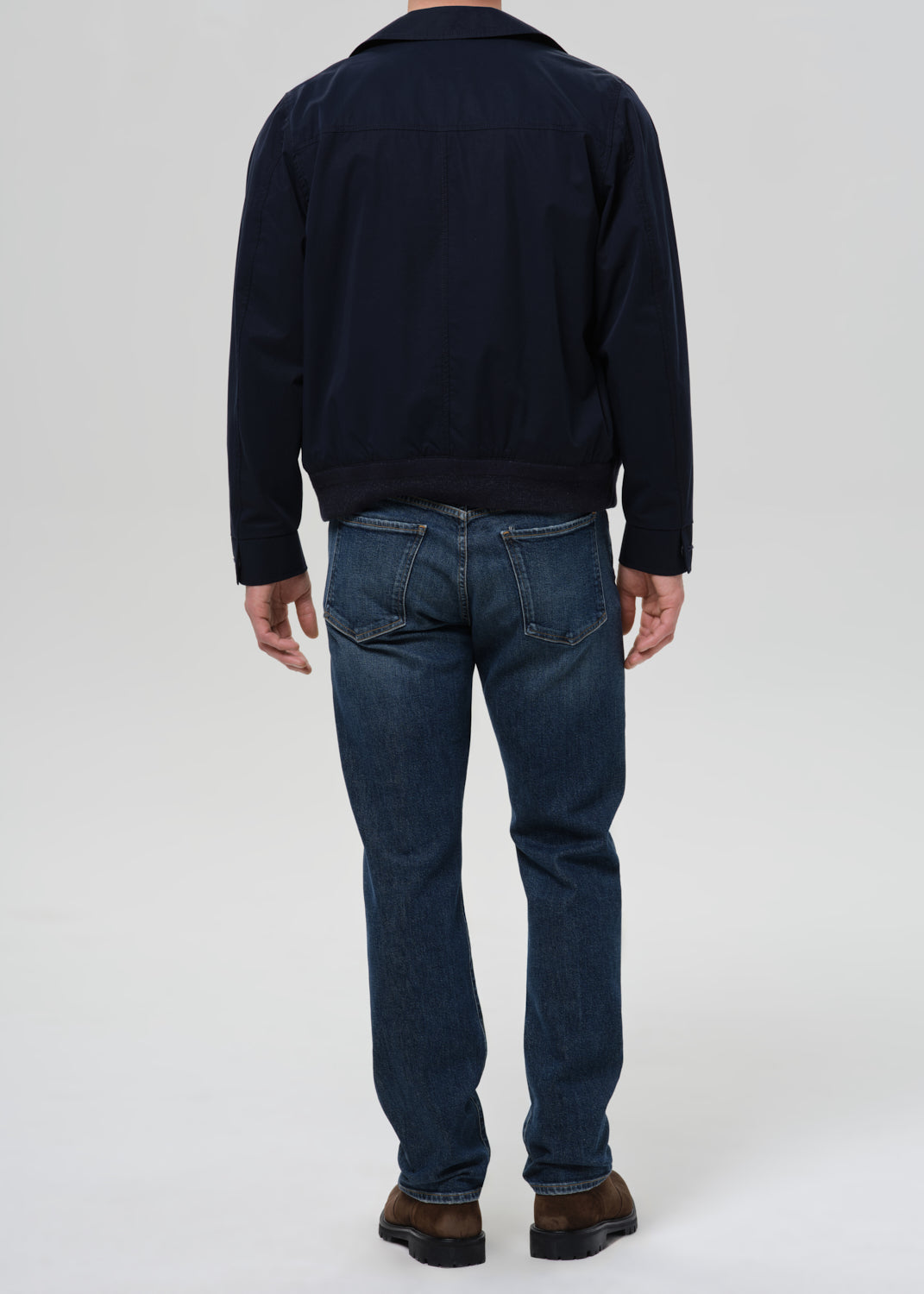 A man viewed from behind wearing a dark navy jacket, dark blue jeans, and brown shoes, standing against a grey background.
