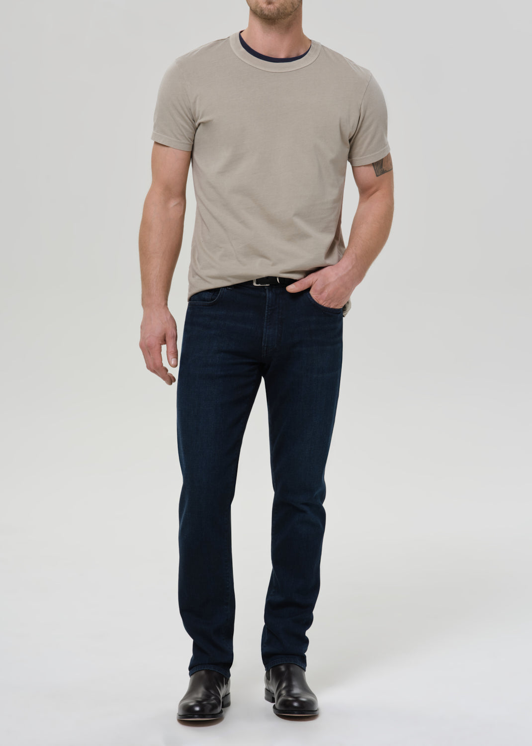 Man wearing a beige t-shirt and dark jeans on a white background
