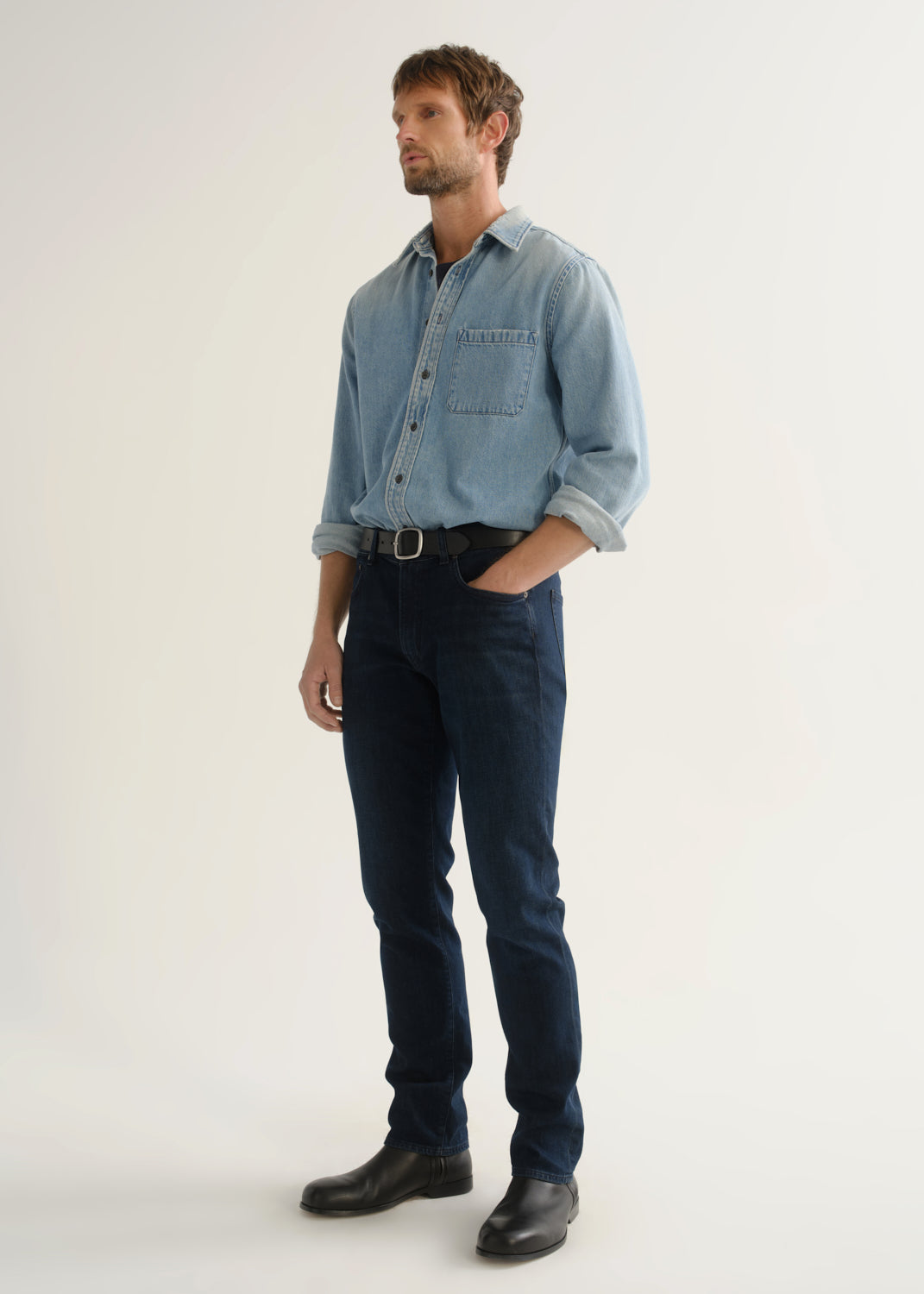 Person wearing a light blue denim shirt, dark indigo jeans, and black boots, front full body view on a light grey background.