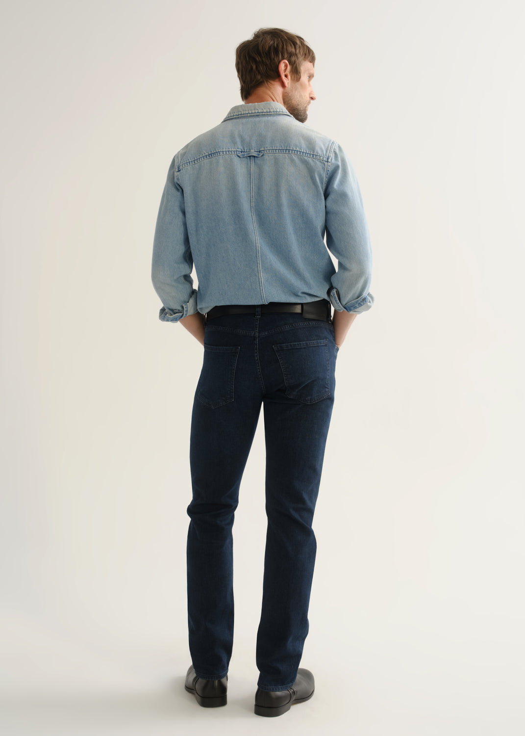 Person wearing a light blue denim shirt, dark indigo jeans, and black boots, back full body view on a light grey background.