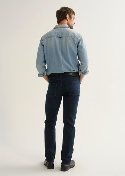 Person wearing a light blue denim shirt, dark indigo jeans, and black boots, back full body view on a light grey background.