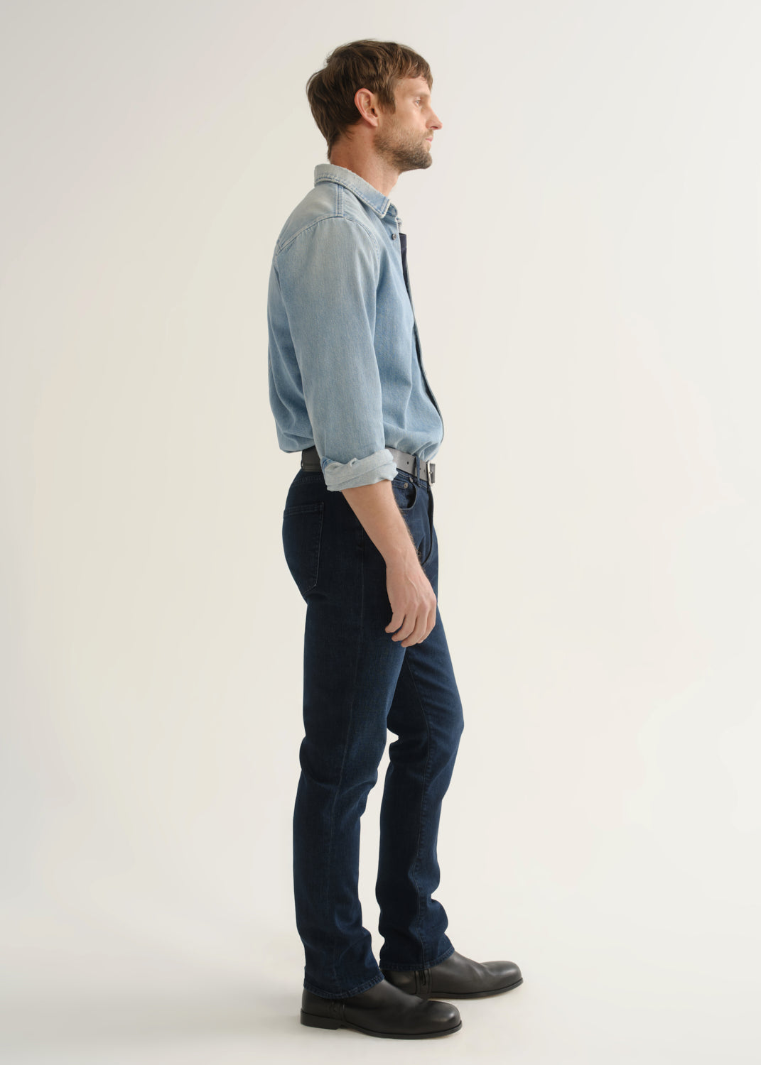 Person wearing a light blue denim shirt, dark indigo jeans, and black boots, side full body view on a light grey background.