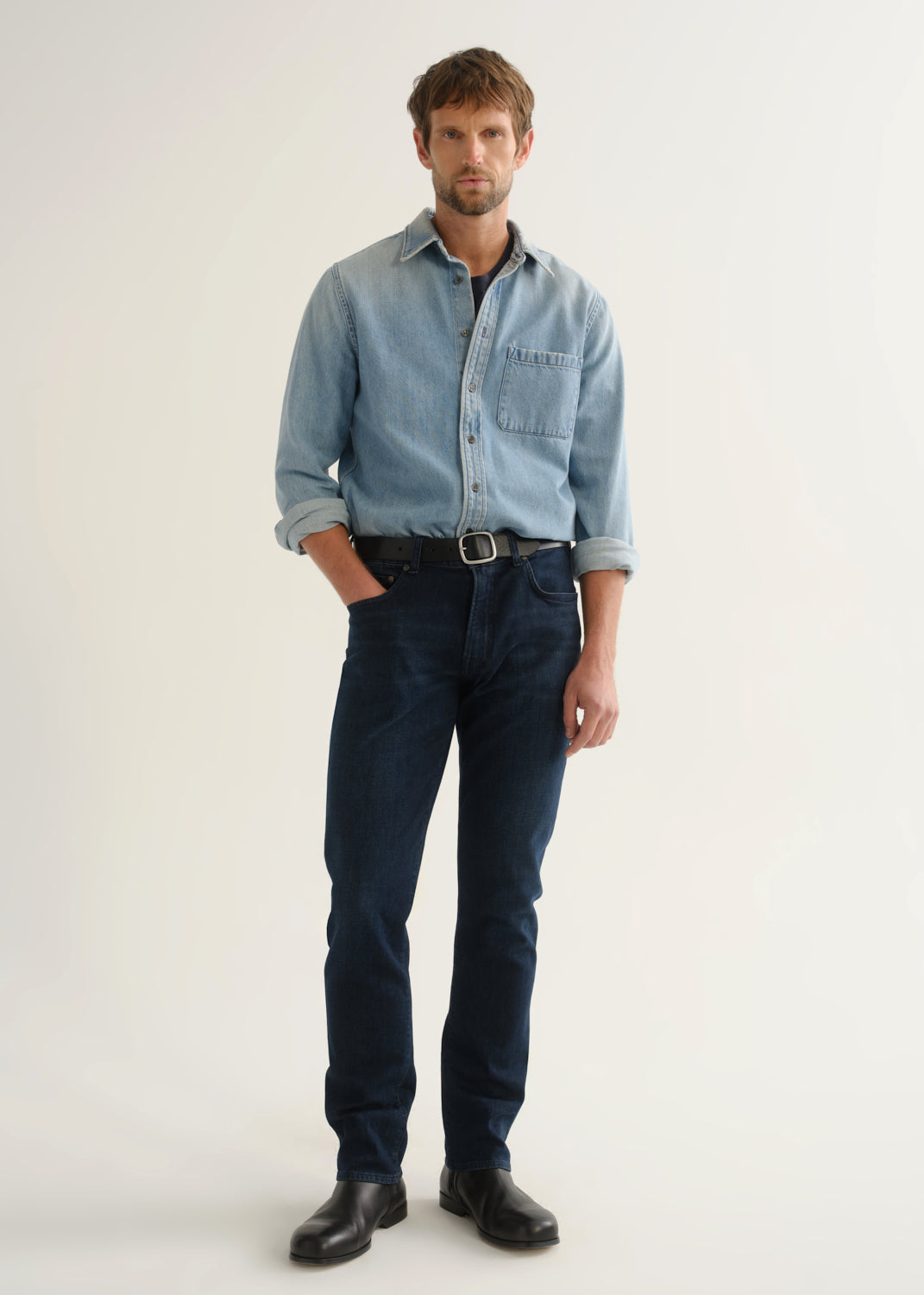 Person wearing a light blue denim shirt, dark indigo jeans, and black boots, angled front full body view on a light grey background.
