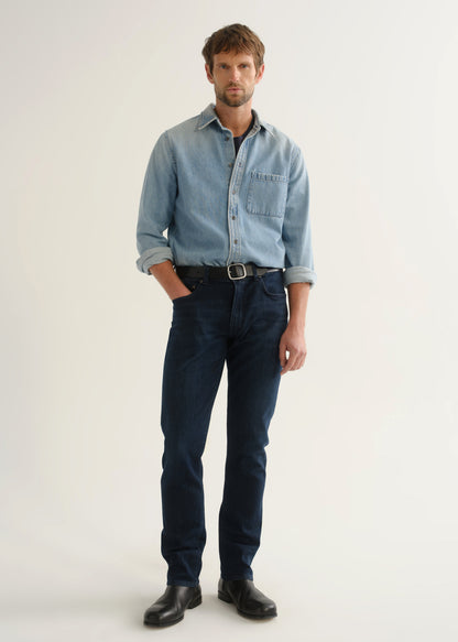 Person wearing a light blue denim shirt, dark indigo jeans, and black boots, angled front full body view on a light grey background.