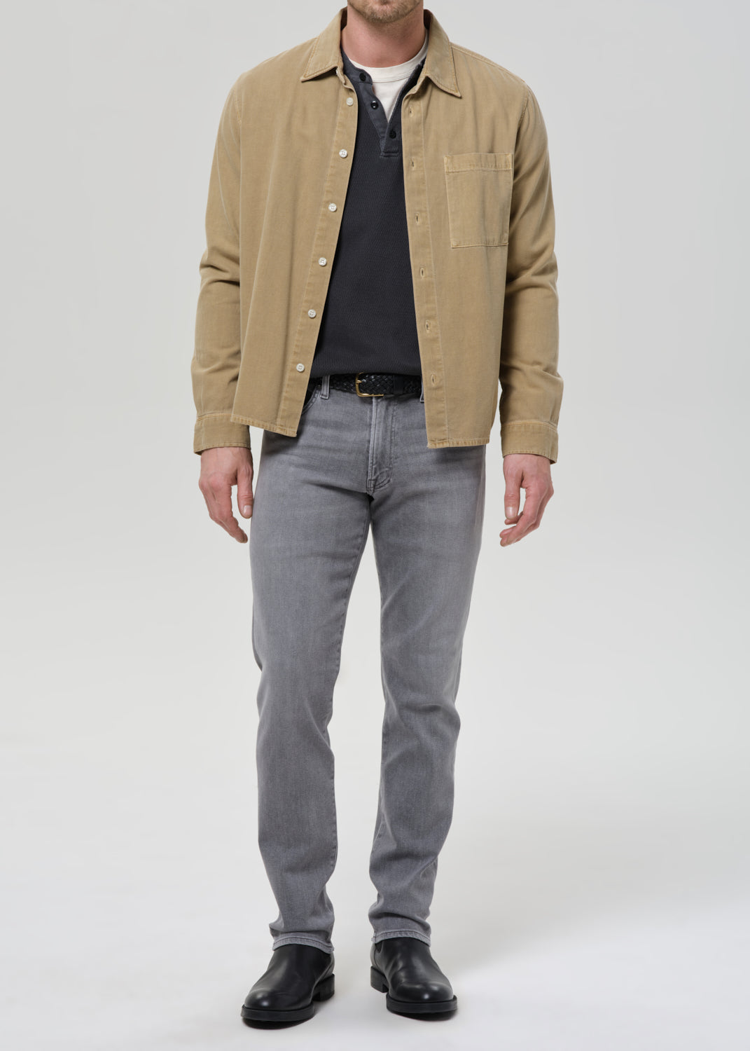 A man wearing a tan overshirt over a dark polo shirt, grey slim-fit jeans, and black leather shoes, standing against a grey background.