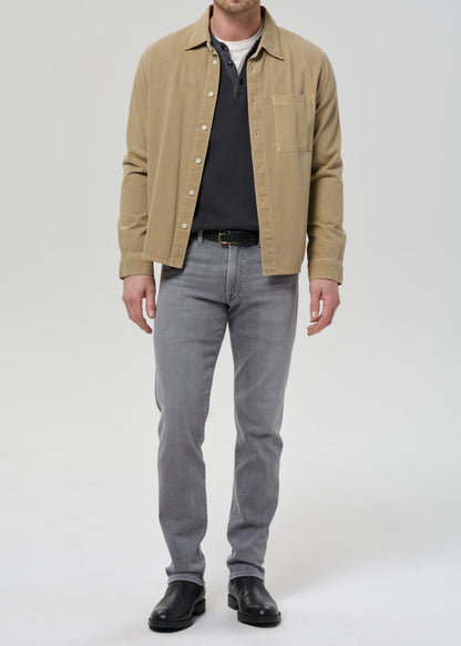 A man wearing a tan overshirt over a dark polo shirt, grey slim-fit jeans, and black leather shoes, standing against a grey background.