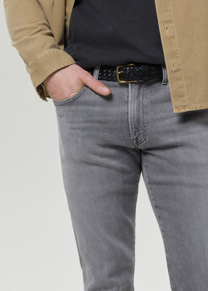 Close-up of a person wearing grey slim-fit jeans, a black braided belt with a gold buckle, a black ribbed top, and a tan overshirt against a grey background.