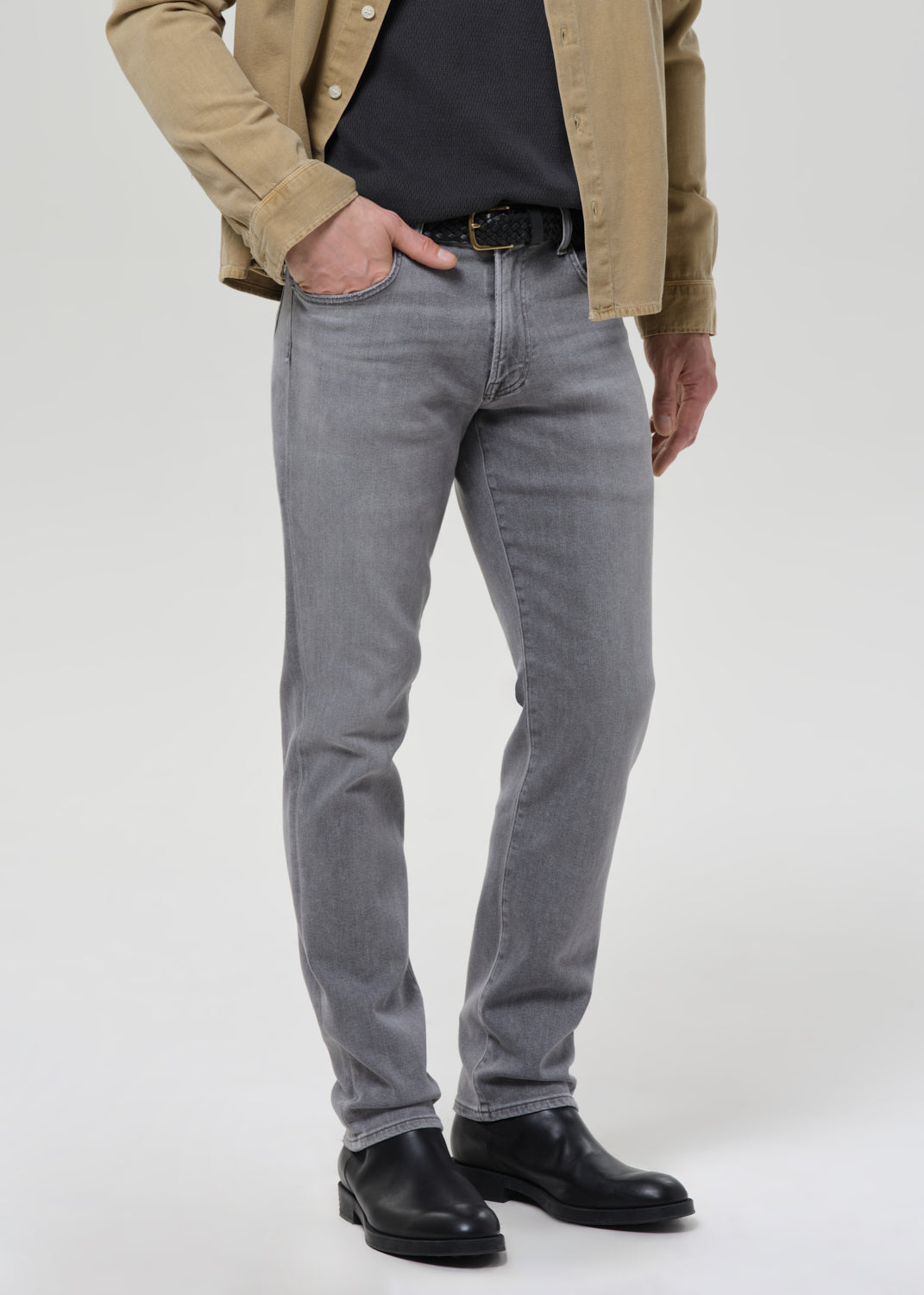 A person wearing slim-fit grey denim jeans, styled with a black shirt, tan overshirt, black belt, and black leather boots, standing against a grey background.