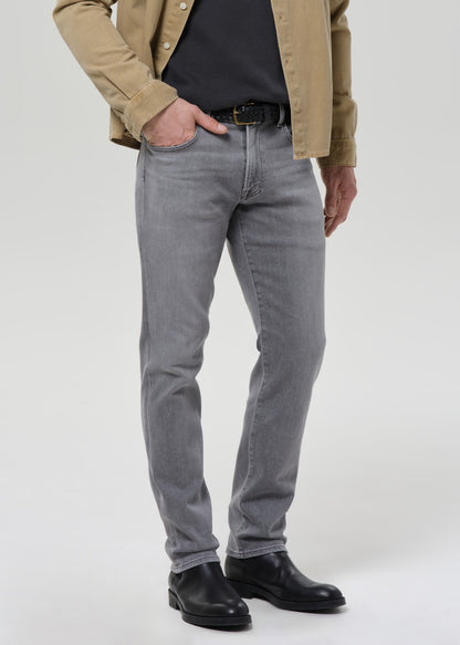 A person wearing slim-fit grey denim jeans, styled with a black shirt, tan overshirt, black belt, and black leather boots, standing against a grey background.
