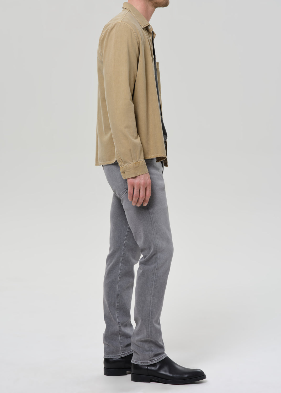 A man shown in profile wearing a beige corduroy shirt jacket, light grey slim-fit jeans, and black leather boots against a grey background.