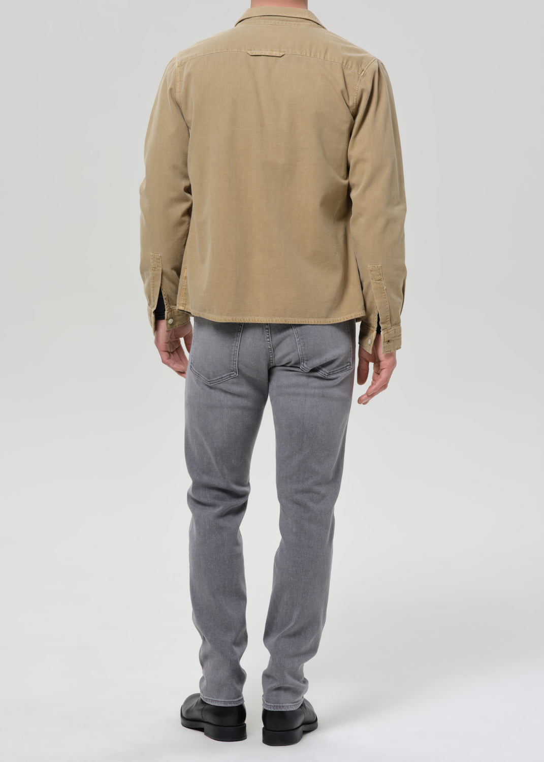 A man viewed from the back wearing a tan long-sleeve button-up shirt and grey slim-fit jeans, paired with black shoes, against a grey background.